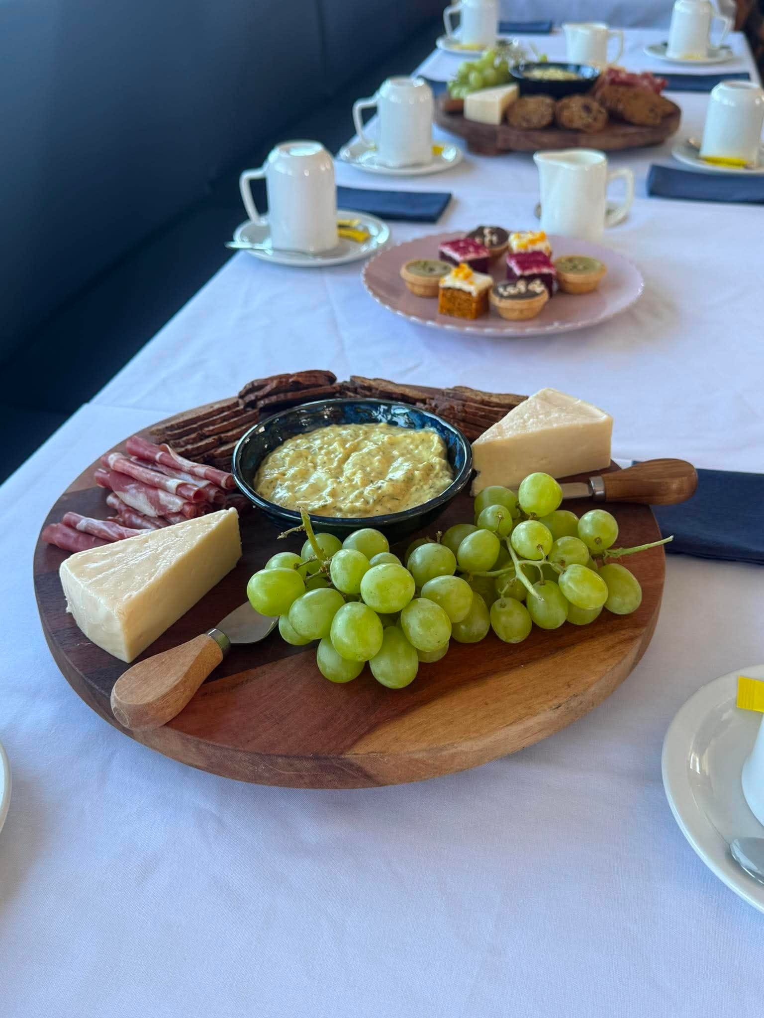A grazing board of cheese, cured meats, dip and grapes on a white tablecloth aboard MV Mini Munya, with petit fours behind